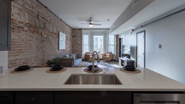 A modern kitchen with a stainless steel sink, a dining area with a sofa and chairs, and a brick wall.