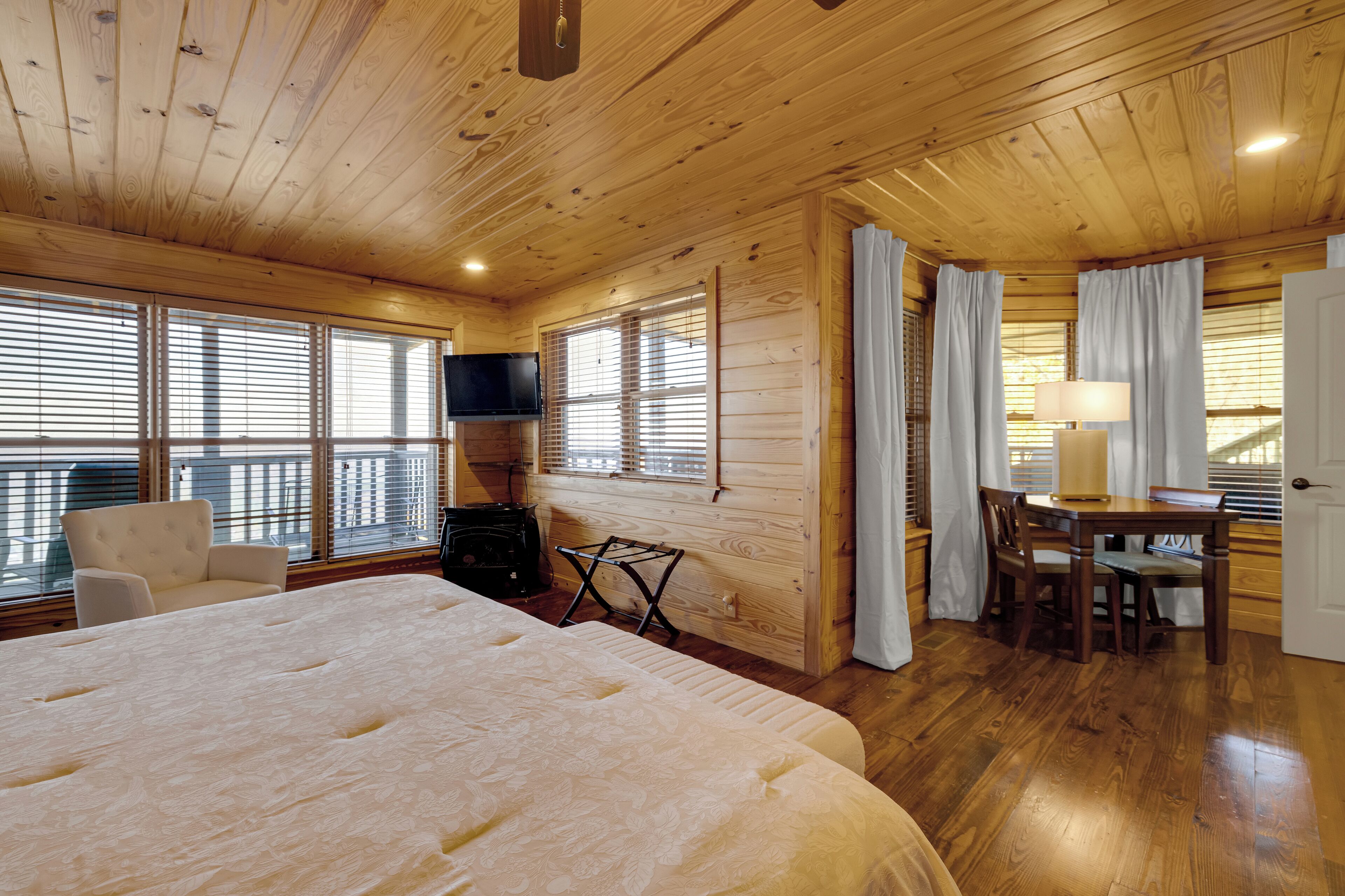 A wooden cabin room with a large bed, a TV, a dining table with chairs, and a balcony with a railing.