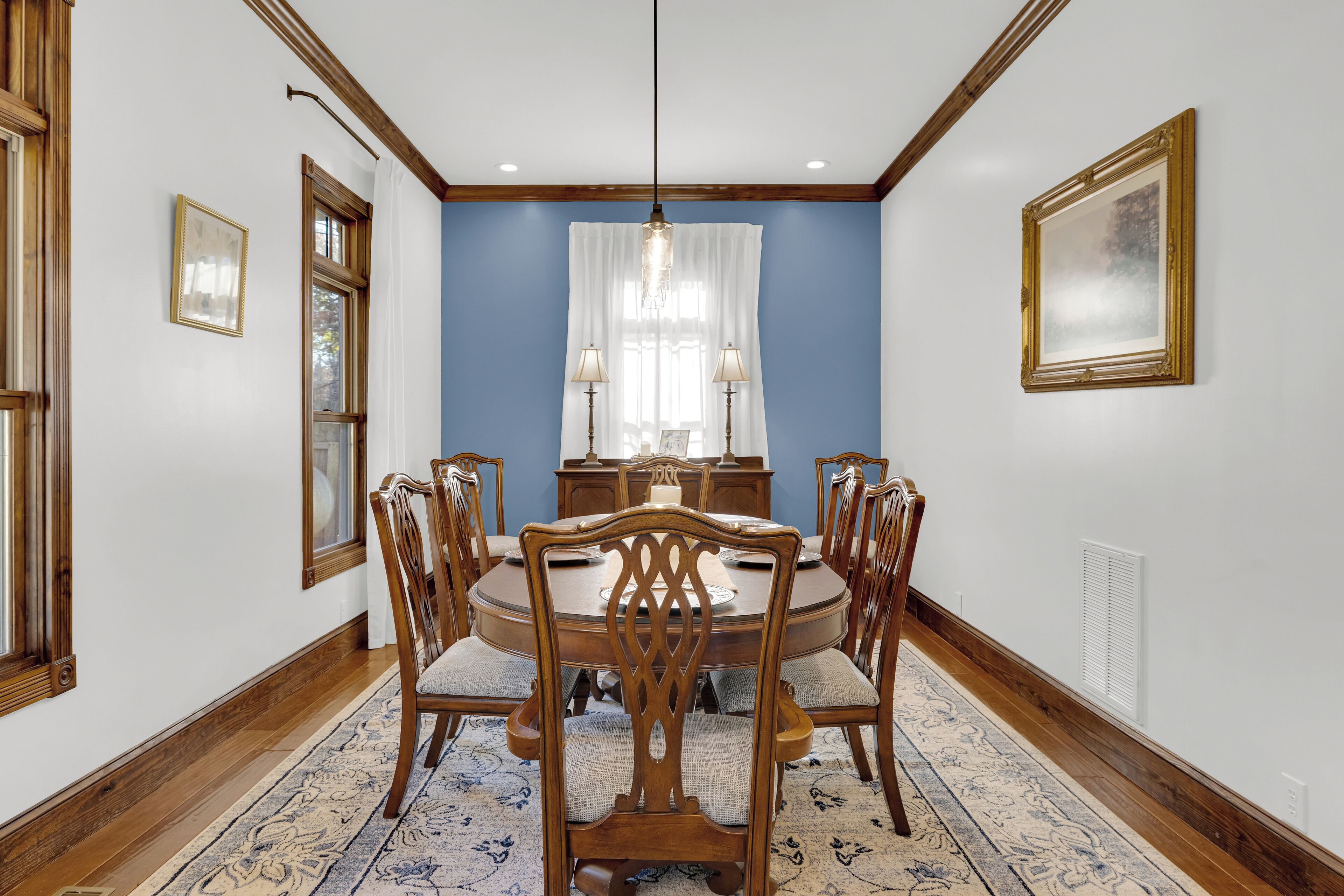 A dining room with a round table, six chairs, a chandelier, and a window with curtains.
