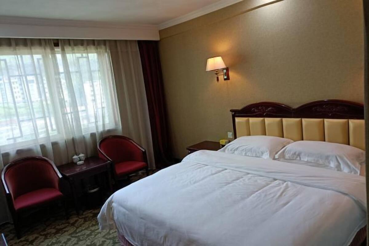 Habitación de hotel con cama, dos sillas rojas, una mesita, una lámpara y una ventana con cortinas.