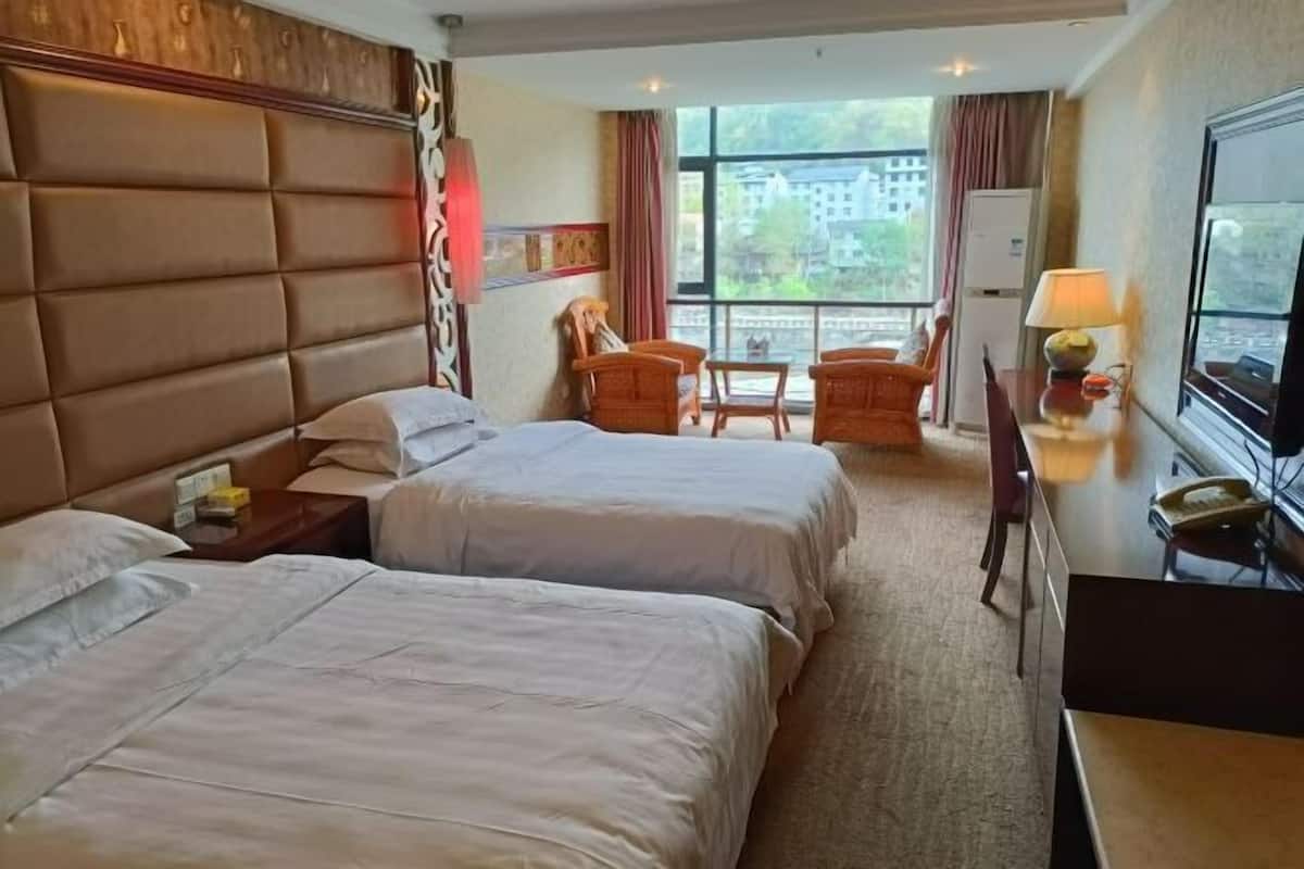 Habitación de hotel con dos camas, un escritorio con lámpara, una silla y vistas a edificios a través de amplios ventanales.