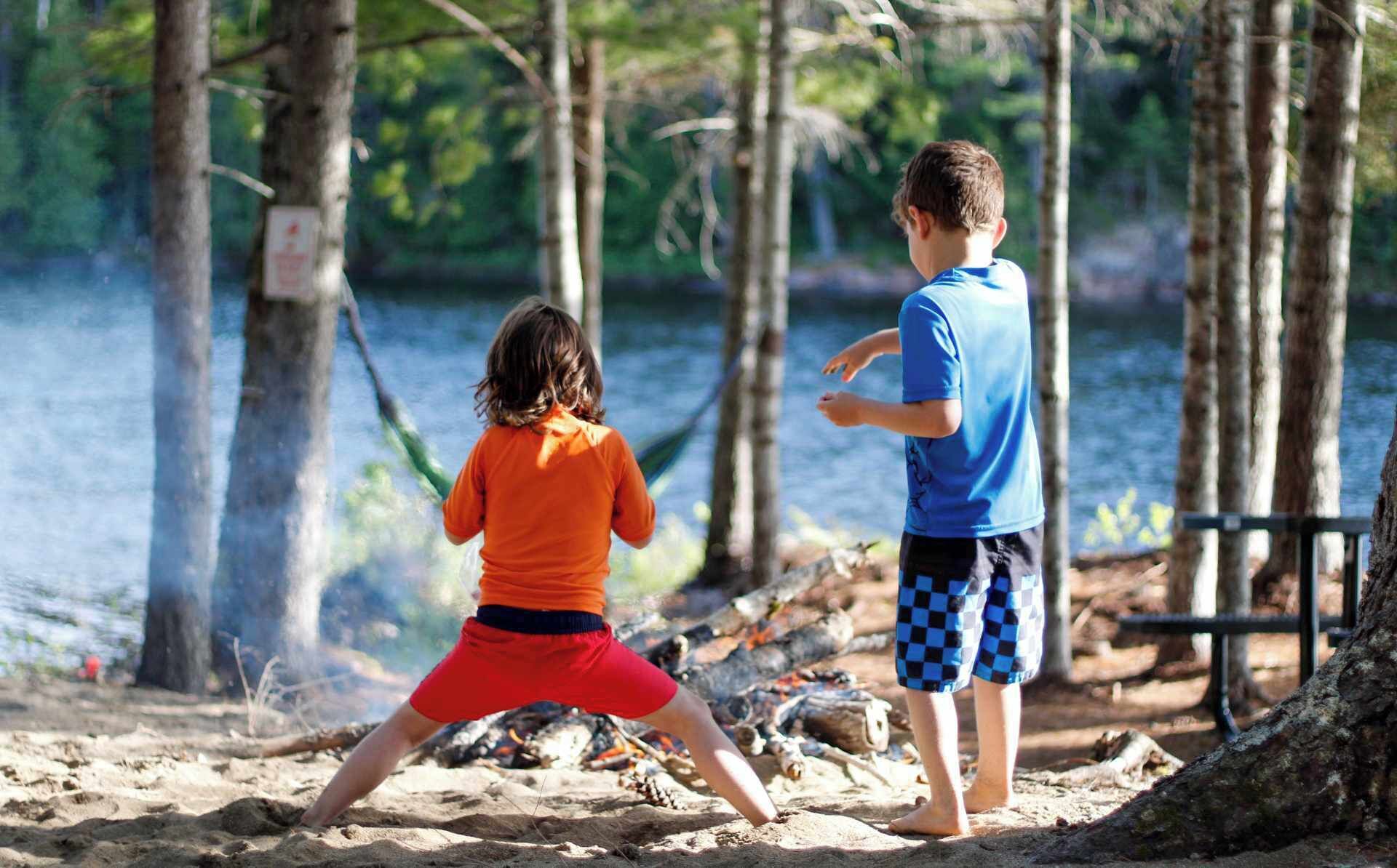 Deux enfants jouent près d’un lac, avec des arbres et un banc en arrière-plan.