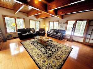 A spacious living room with a wooden ceiling, large windows, a black leather sofa, a coffee table, and a patterned rug. - Bear Mountain Lodge (Robbinston)