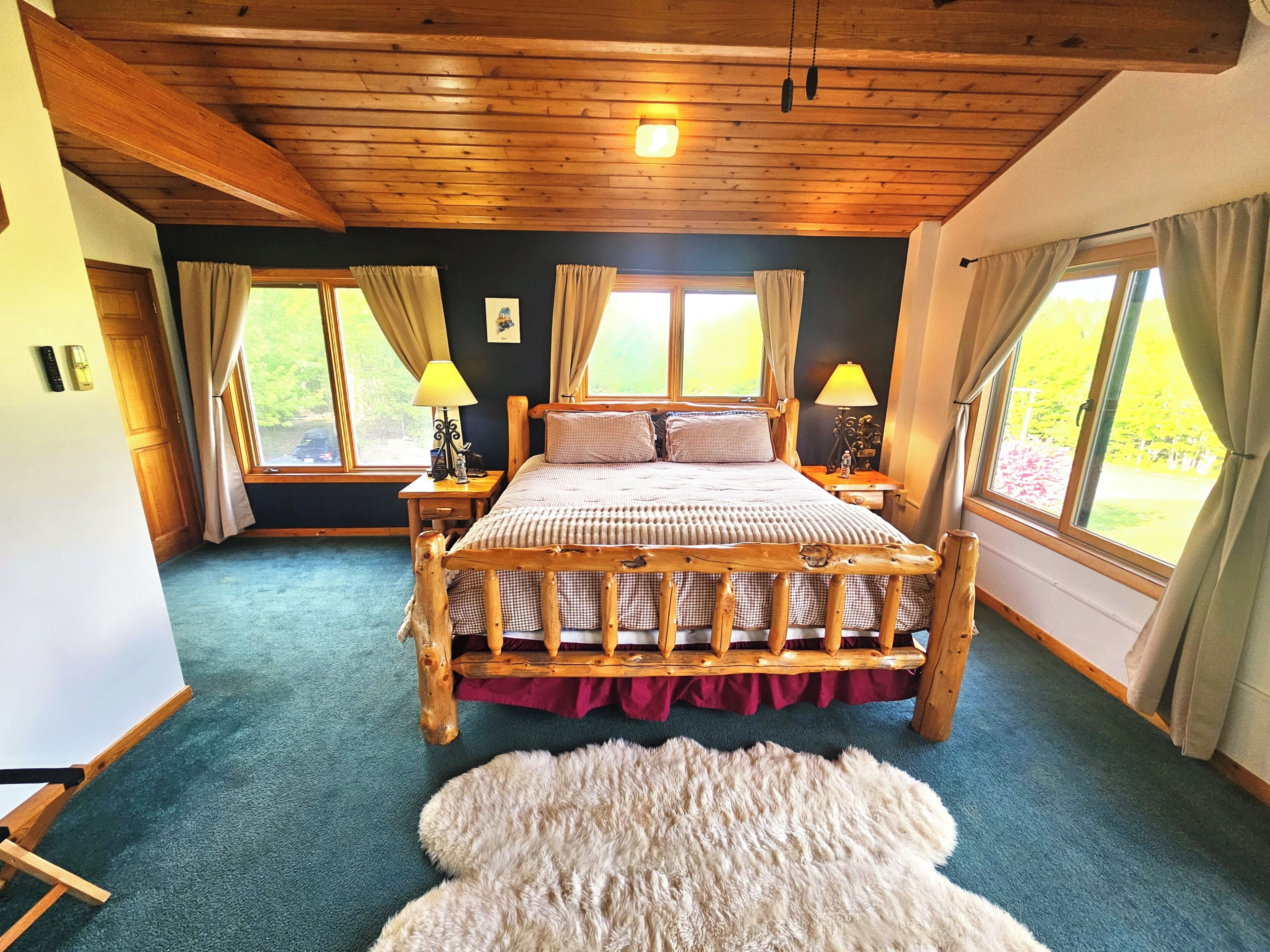 Una cama con estructura de madera, falda roja, dos lámparas de noche y vista a la vegetación a través de ventanas.
