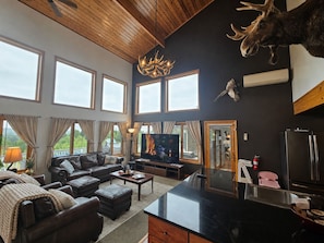 A spacious living room with a fireplace, a mounted moose head, and a large window overlooking a scenic view. - Bear Mountain Lodge (Robbinston)