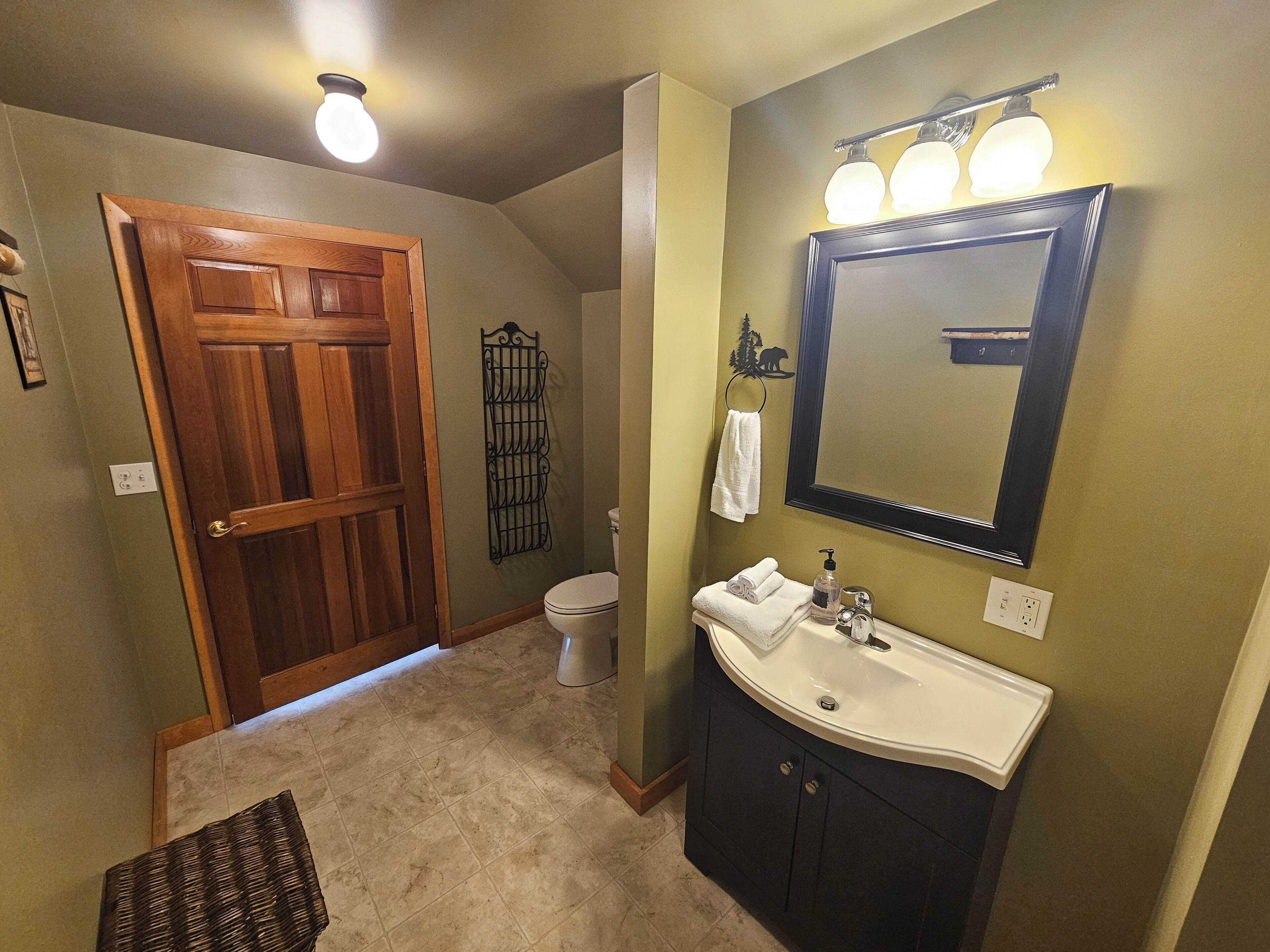Un baño con puerta de madera, inodoro, lavabo con espejo y toallero.
