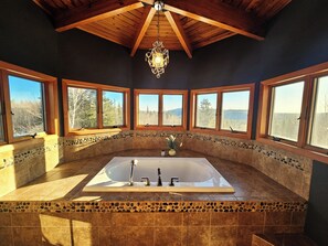 A bathroom with a large bathtub, stone mosaic border, wooden beams, and large windows with a mountain view. - Bear Mountain Lodge (Robbinston)