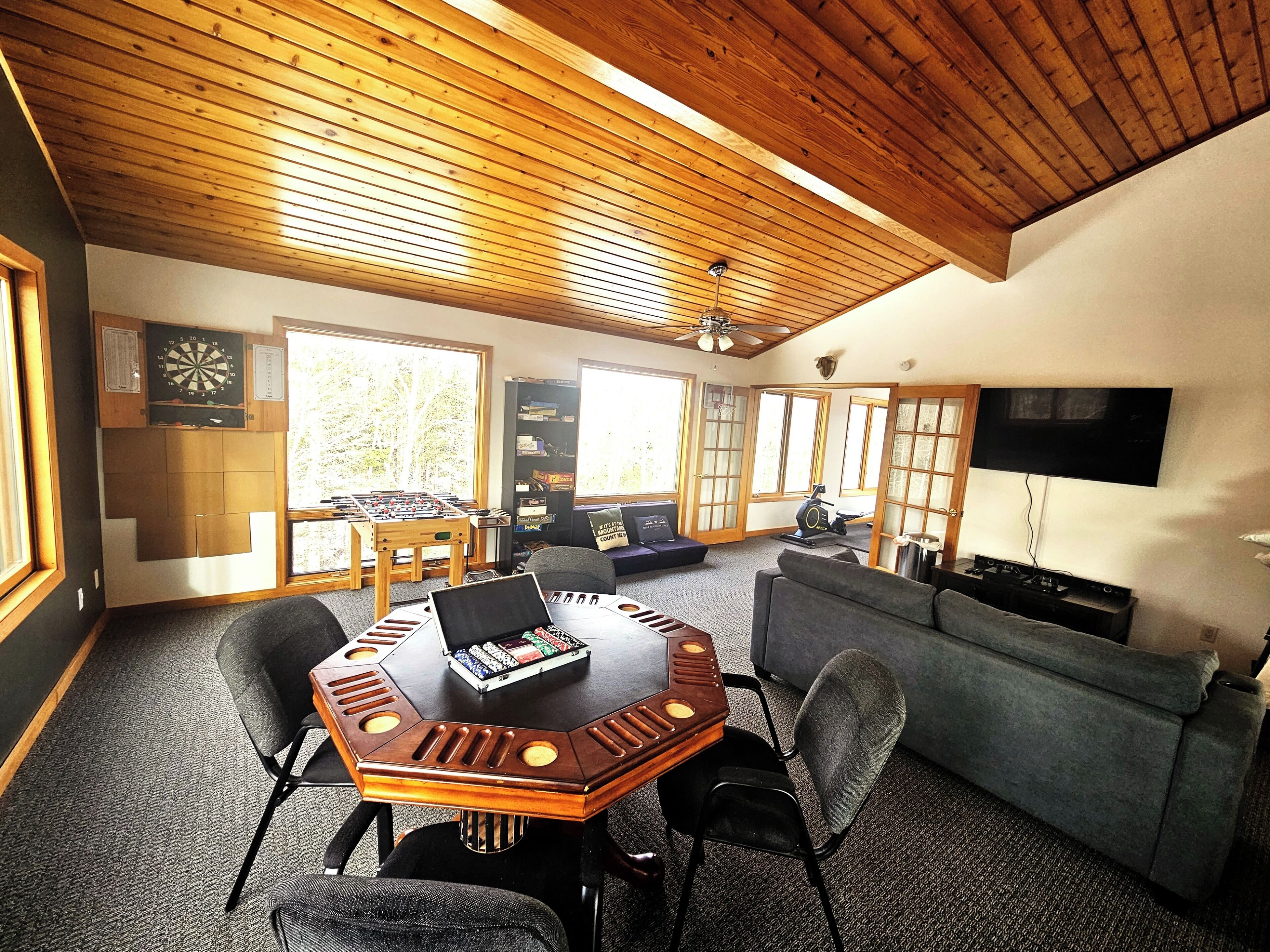 A spacious living room with a wooden ceiling, a large window, a television, a foosball table, and a treadmill.