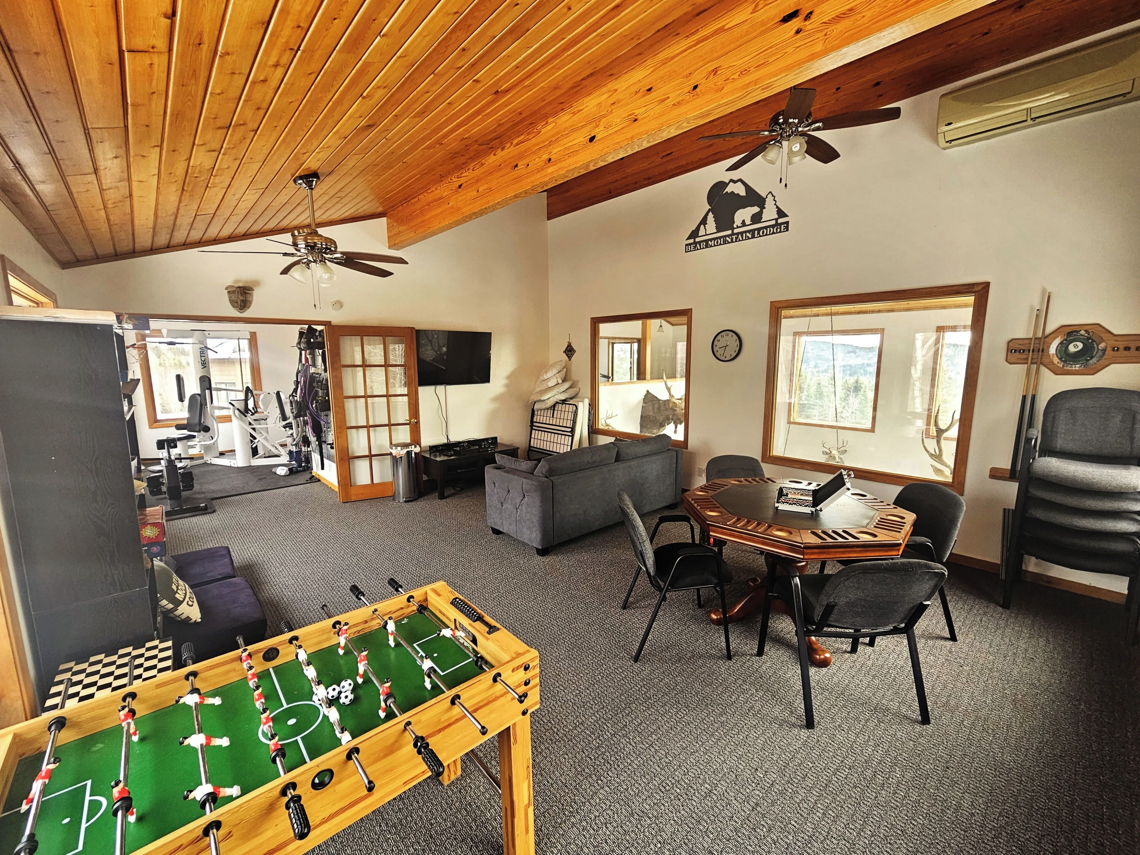 A cozy lodge interior with a foosball table, a dining area, and a gym visible through an open door.