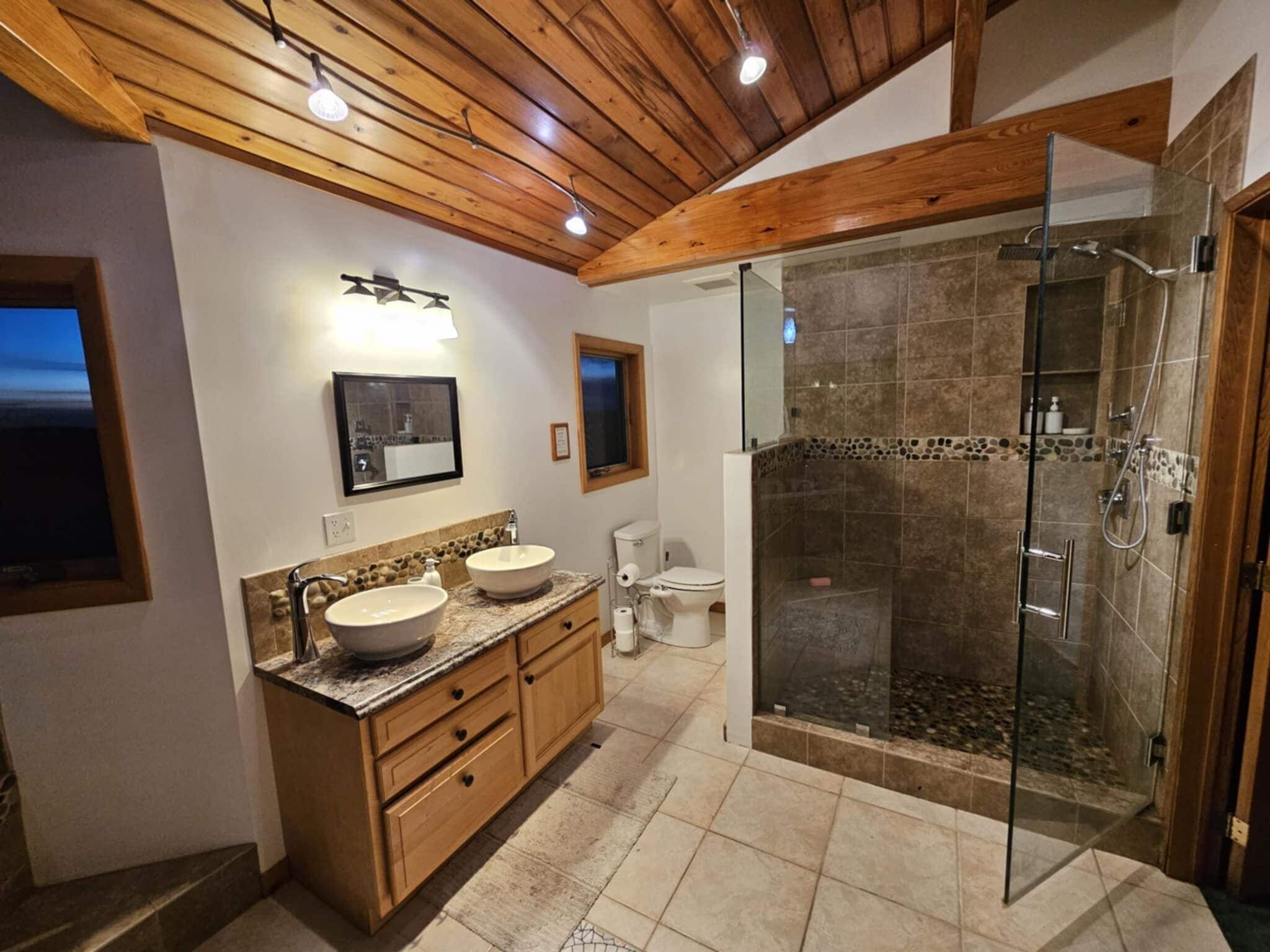 A modern bathroom with a double sink vanity, a glass-enclosed shower, and a wooden ceiling.
