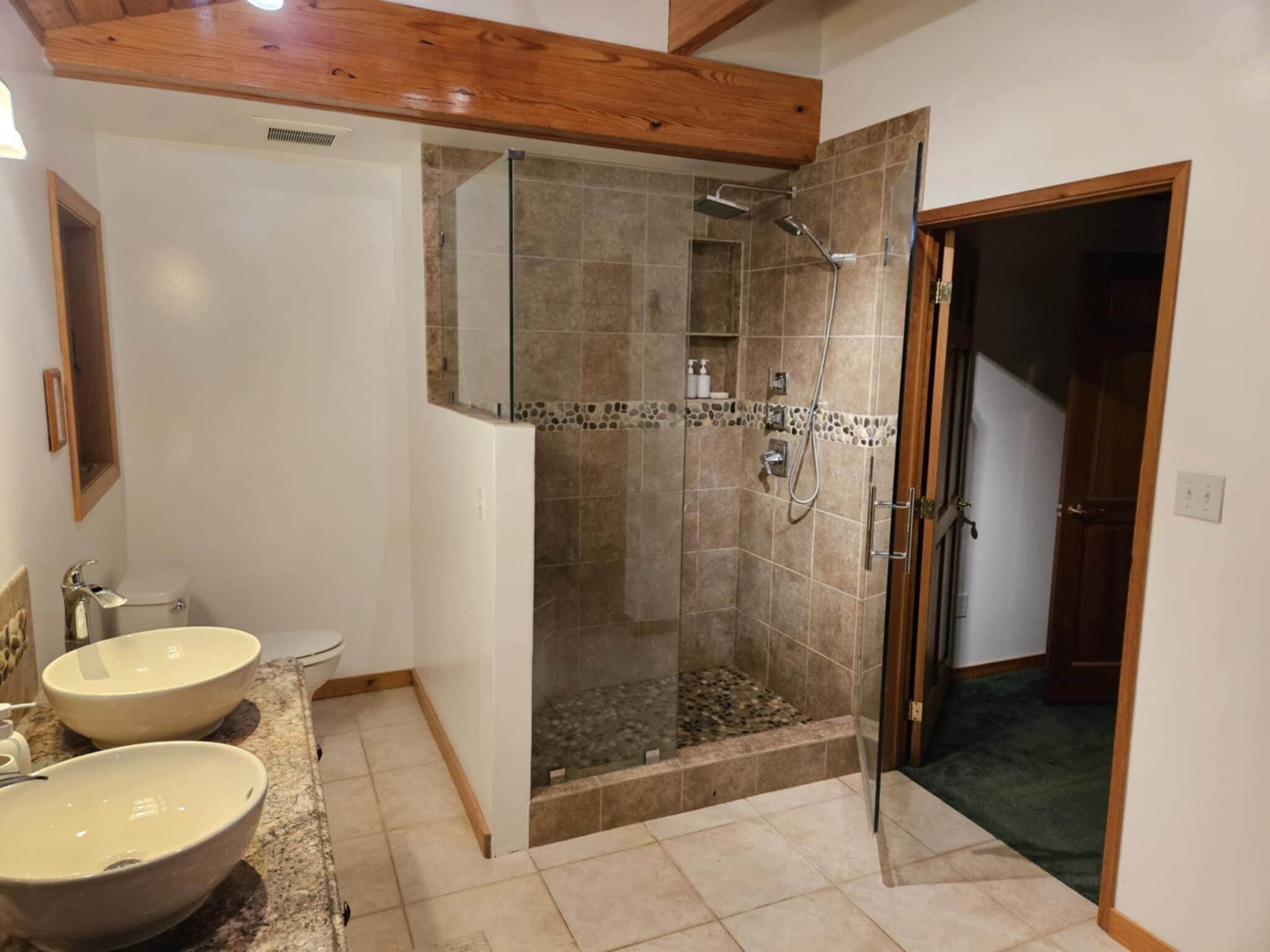 Un baño con dos lavamanos, ducha y puerta que da a otra habitación.
