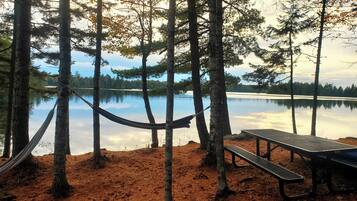 A hammock tied between trees, a picnic table, and a calm lake with reflections.