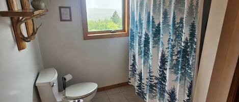 A bathroom with a toilet, a window, a shower curtain with a forest pattern, a wooden shelf with plants, and a tiled floor.