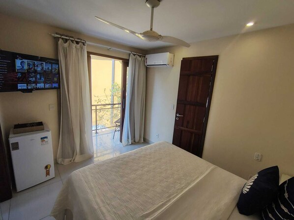 Une chambre à coucher comprenant un lit, un téléviseur fixé au mur, un ventilateur de plafond et une porte donnant sur un balcon.