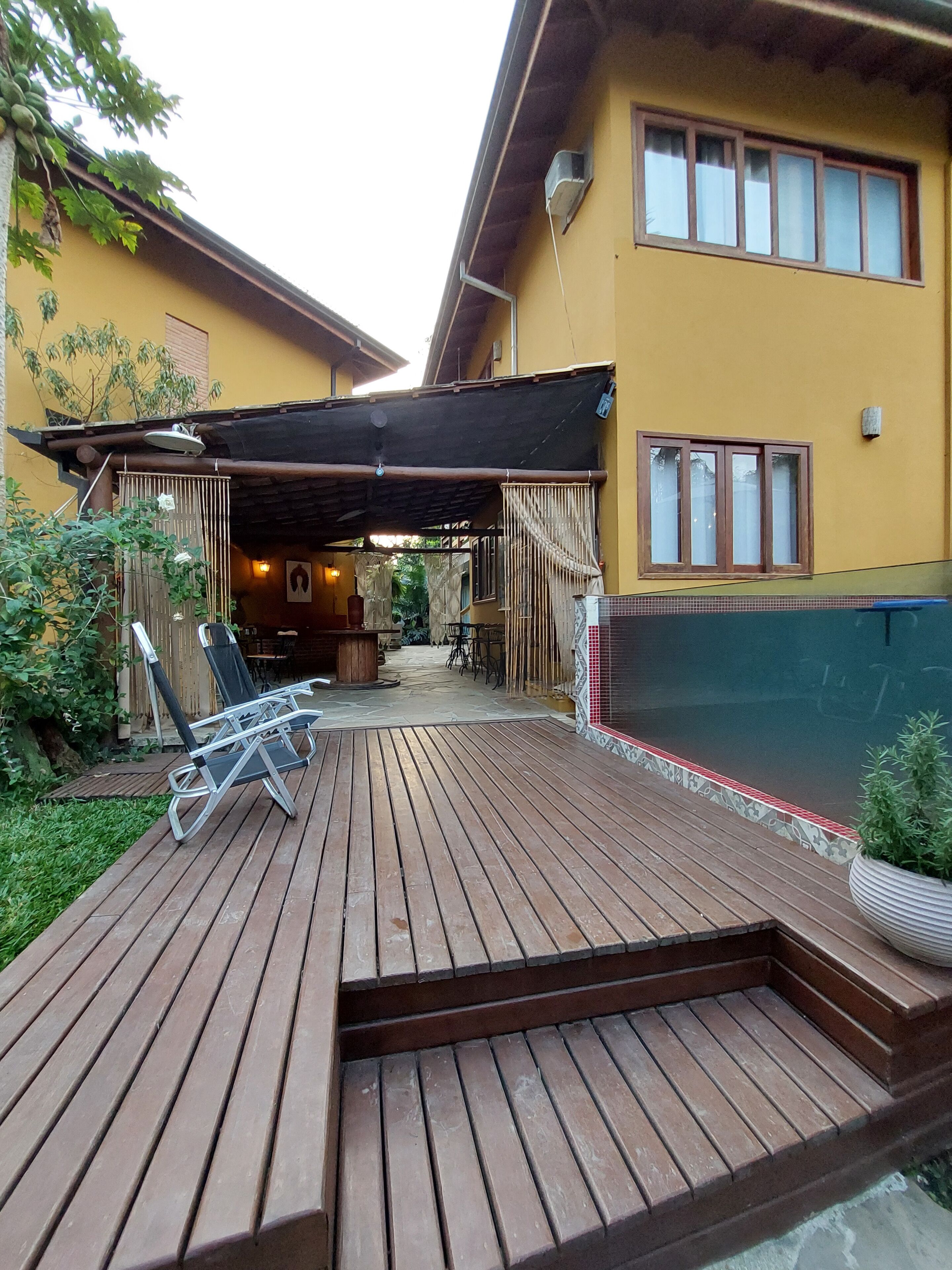 Una terraza de madera con sillones de descanso y una zona cubierta para sentarse, junto a un edificio amarillo con piscina.
