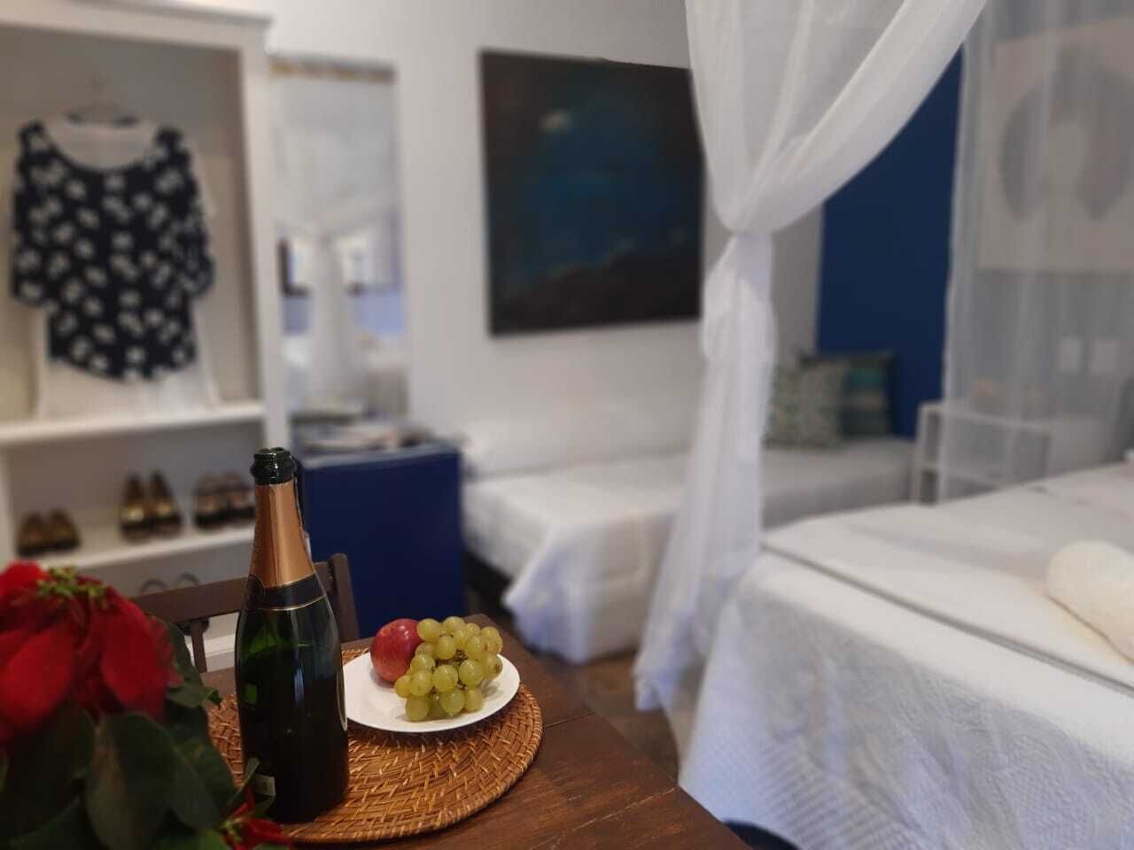 Habitación de hotel con una cama, una mesita de noche con una botella y uvas, una pequeña mesa con un plato de uvas, una rosa roja y una silla azul.