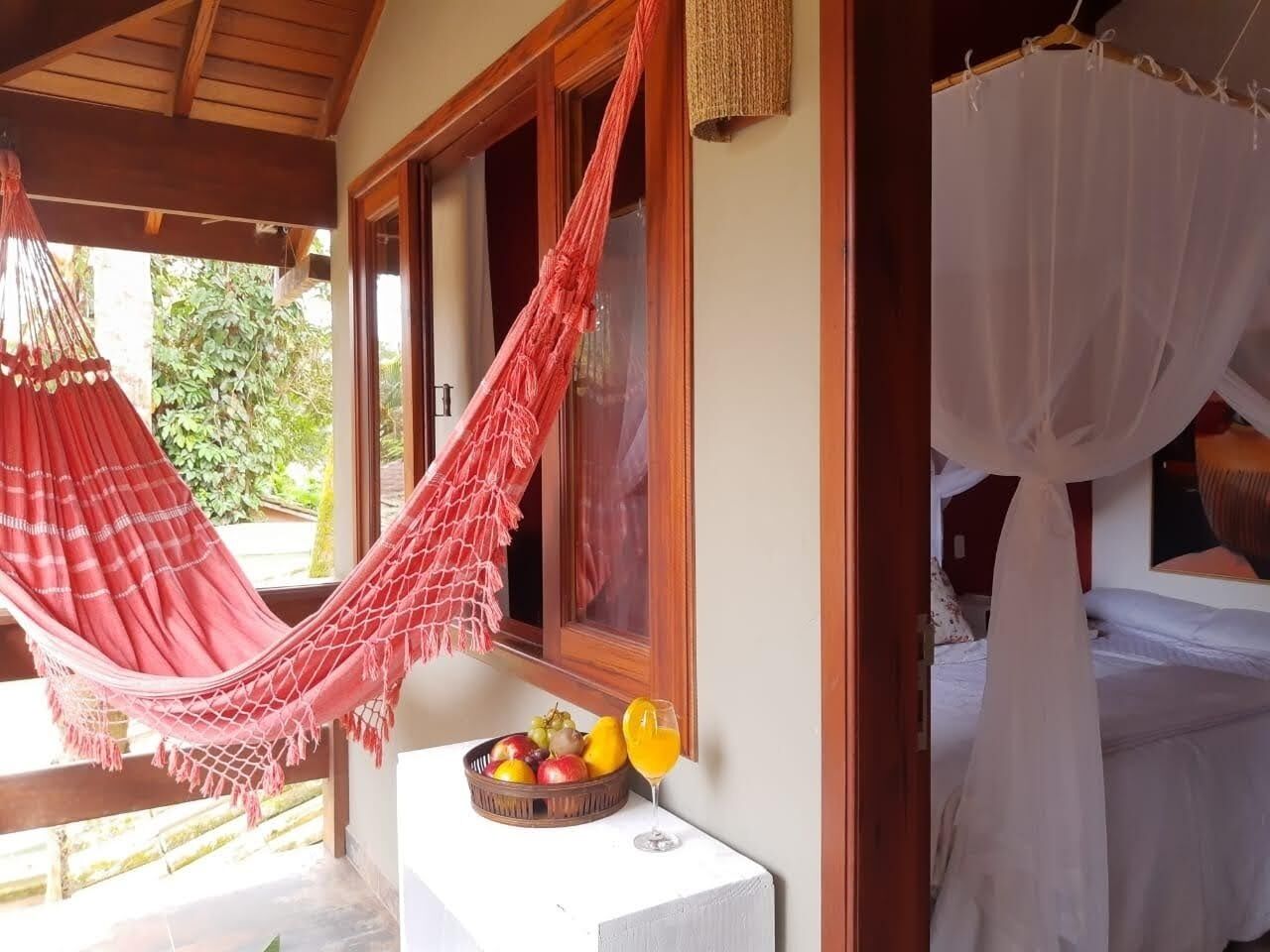 A room with a hammock, a bed with a canopy, and a fruit bowl with a drink on a table.