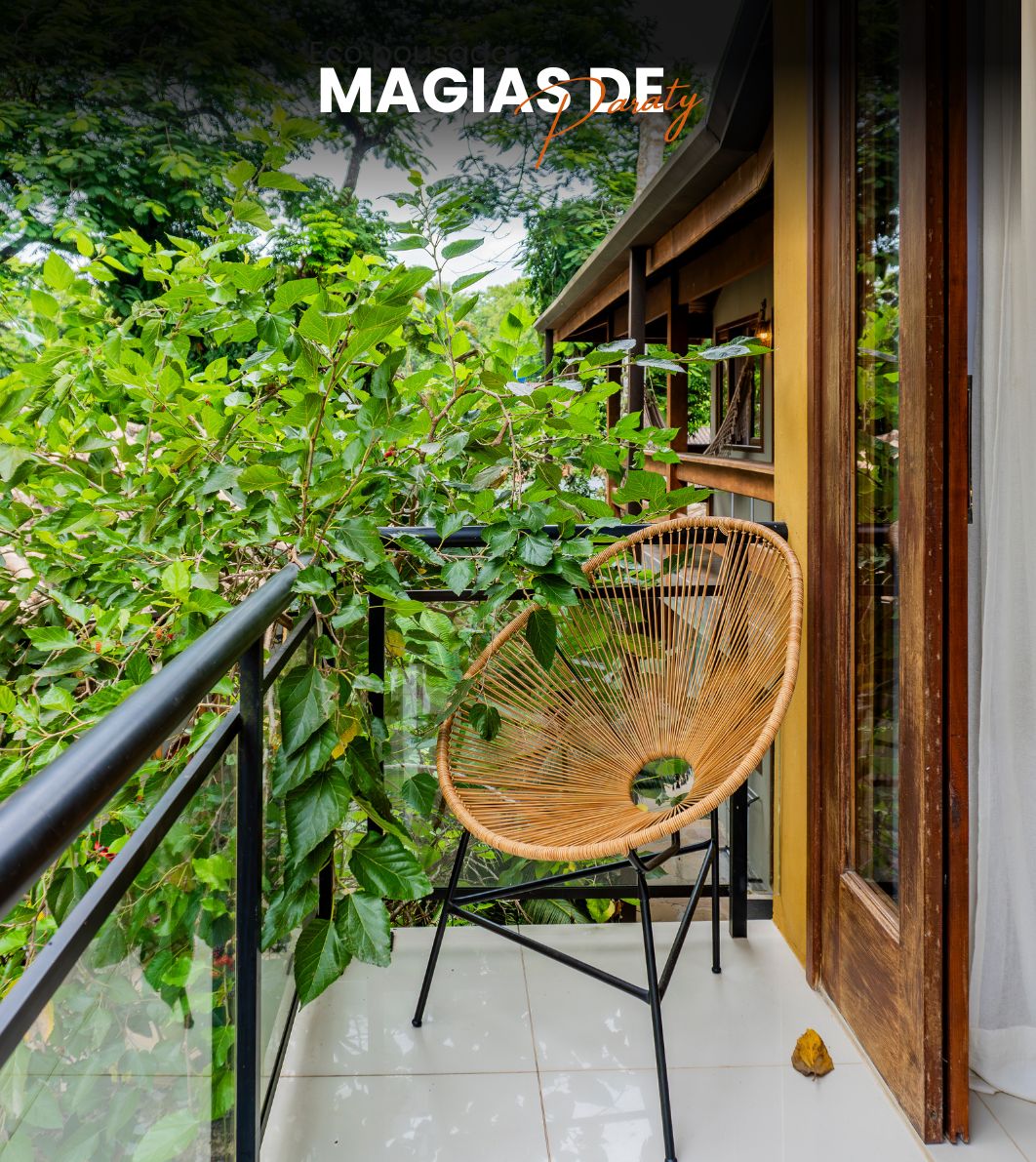 Un balcón con una silla de mimbre, abundante vegetación y barandilla de madera.