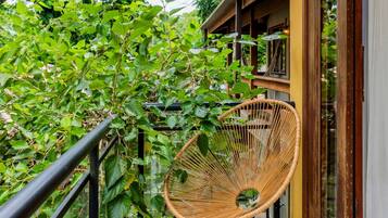 Un balcón con una silla de mimbre, abundante vegetación y barandilla de madera.