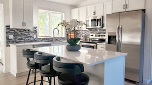 A modern kitchen with white cabinets, a stainless steel refrigerator, a built-in oven, and a large island with bar stools. - Modern Home with Game Room, Private Yard, Panoramic Views, 5B Sleeps 8+ (Spokane)