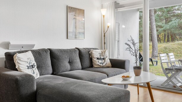 A modern living room with a grey sofa, a small round table with a bowl of fruit, a potted plant, and a view of trees through a glass door. - 1 bedroom gorgeous apartment in Nexø (Nexø)