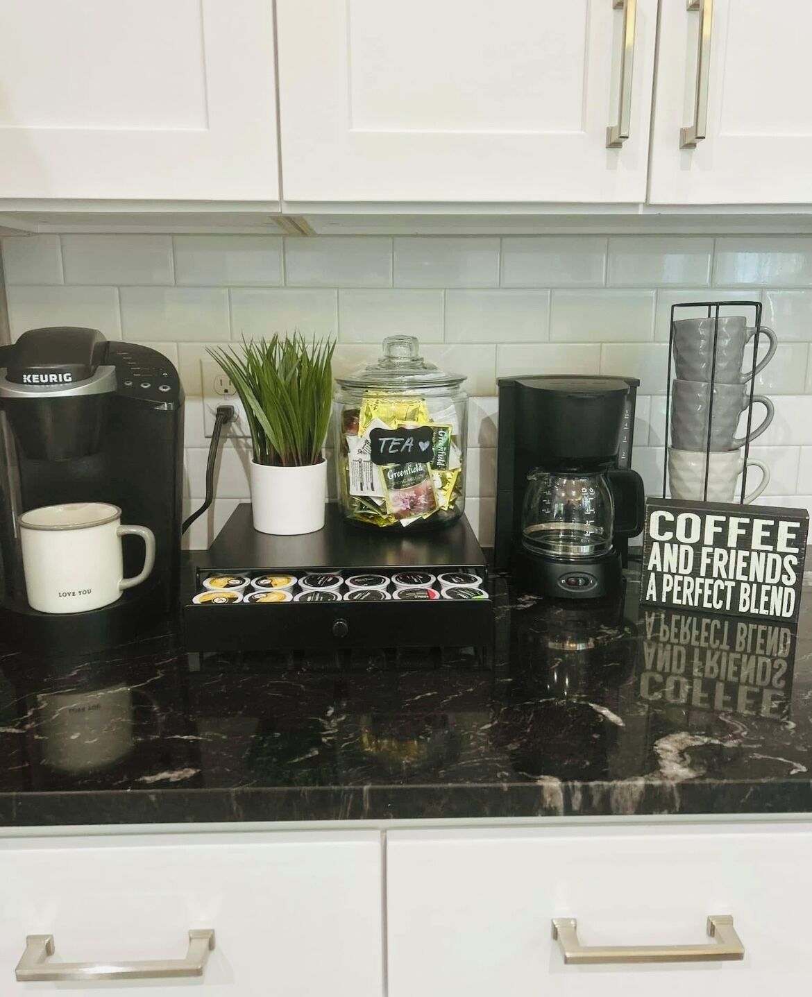 Un comptoir de cuisine avec une machine à café Keurig, une tasse, un bocal de thé, une autre machine à café et un panneau sur lequel on peut lire : « Le café et les amis, un mélange parfait ».