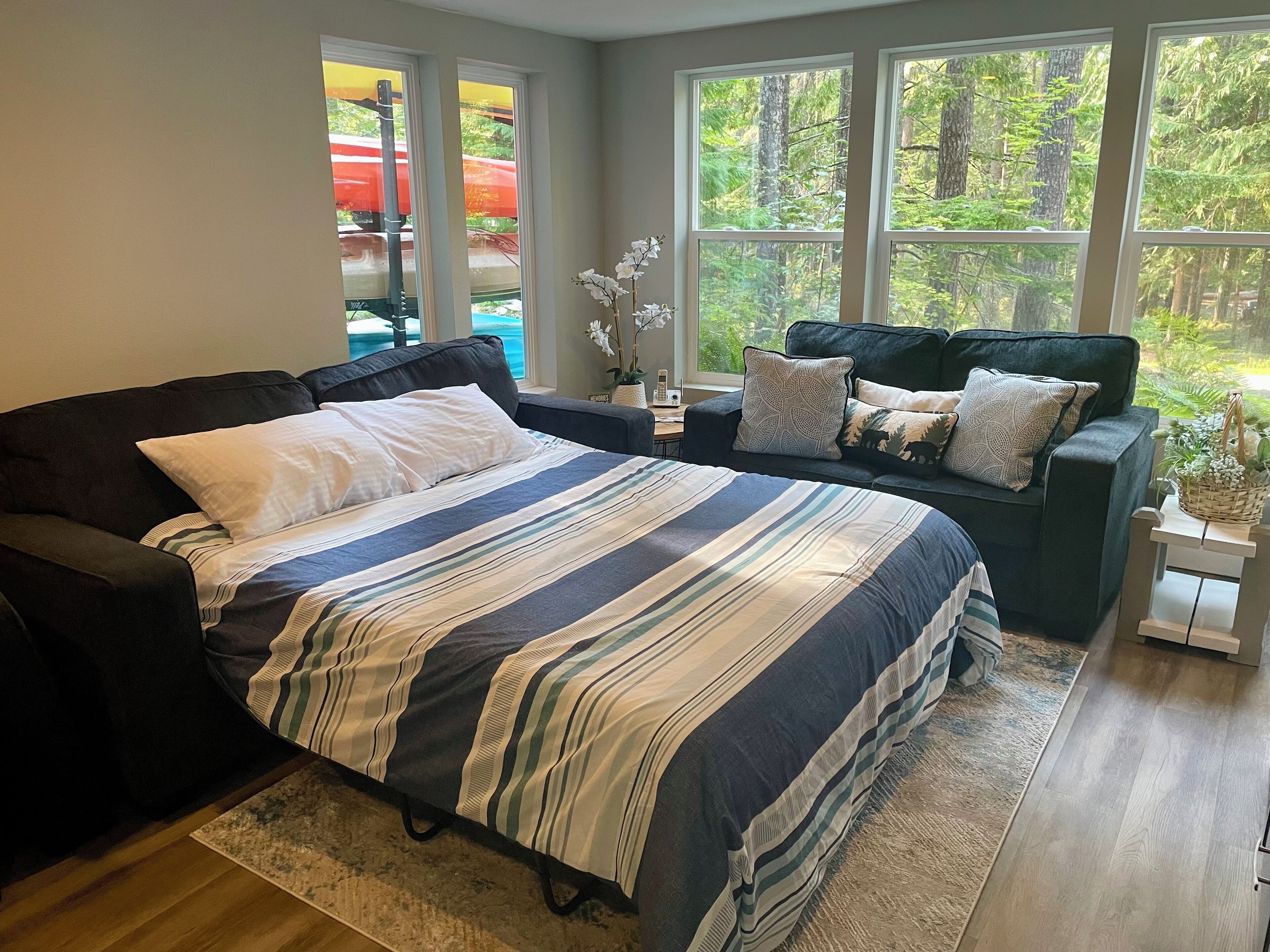 A bedroom with a large bed, a dark sofa, and a view of a forest through large windows.