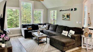 Ein modernes Wohnzimmer mit einer Eckcouch, einem Couchtisch und großen Fenstern mit Blick auf Bäume.