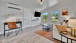 Un salon moderne avec un plancher en bois, un téléviseur fixé au mur, un bureau avec une chaise, un canapé avec une lampe et une grande fenêtre donnant sur l’extérieur.