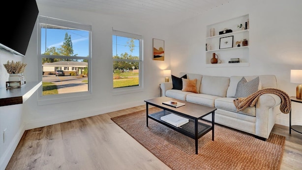 Un salon moderne avec un canapé blanc, une table basse et un téléviseur fixé au mur. Deux fenêtres offrent une vue sur l’extérieur.