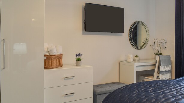 A bedroom with a bed, a dresser with drawers, a vanity with a mirror, and a television on the wall.
