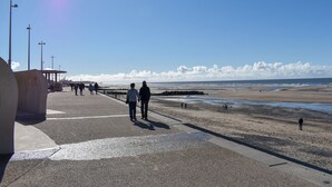 A coastal walkway with a clear sky, a few people walking, and a distant view of the sea.