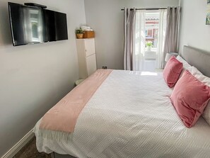 Un dormitorio con cama, televisión montada en la pared, ventana con cortinas y un mueble pequeño.