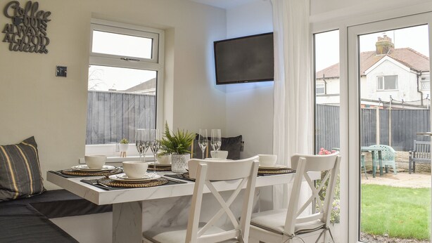 Uma sala de jantar com uma mesa posta para quatro pessoas, um sofá e uma TV fixada na parede.