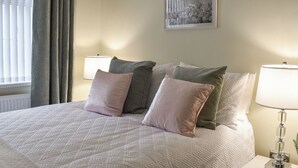 Cama bem arrumada com colcha estampada e várias almofadas, abajur de cabeceira e quadro na parede.