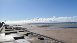 Uma praia com um calçadão de concreto que leva até a orla, céu claro e o oceano ao fundo.