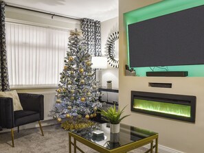A living room with a Christmas tree, a black sofa, a glass coffee table, a flat-screen TV, and a fireplace.