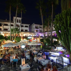 An outdoor dining area with tables and chairs, palm trees, and a building with multiple floors in the background. - Orihuela Costa, Sleeps 6 With Roof Terrace & Pool (Orihuela)