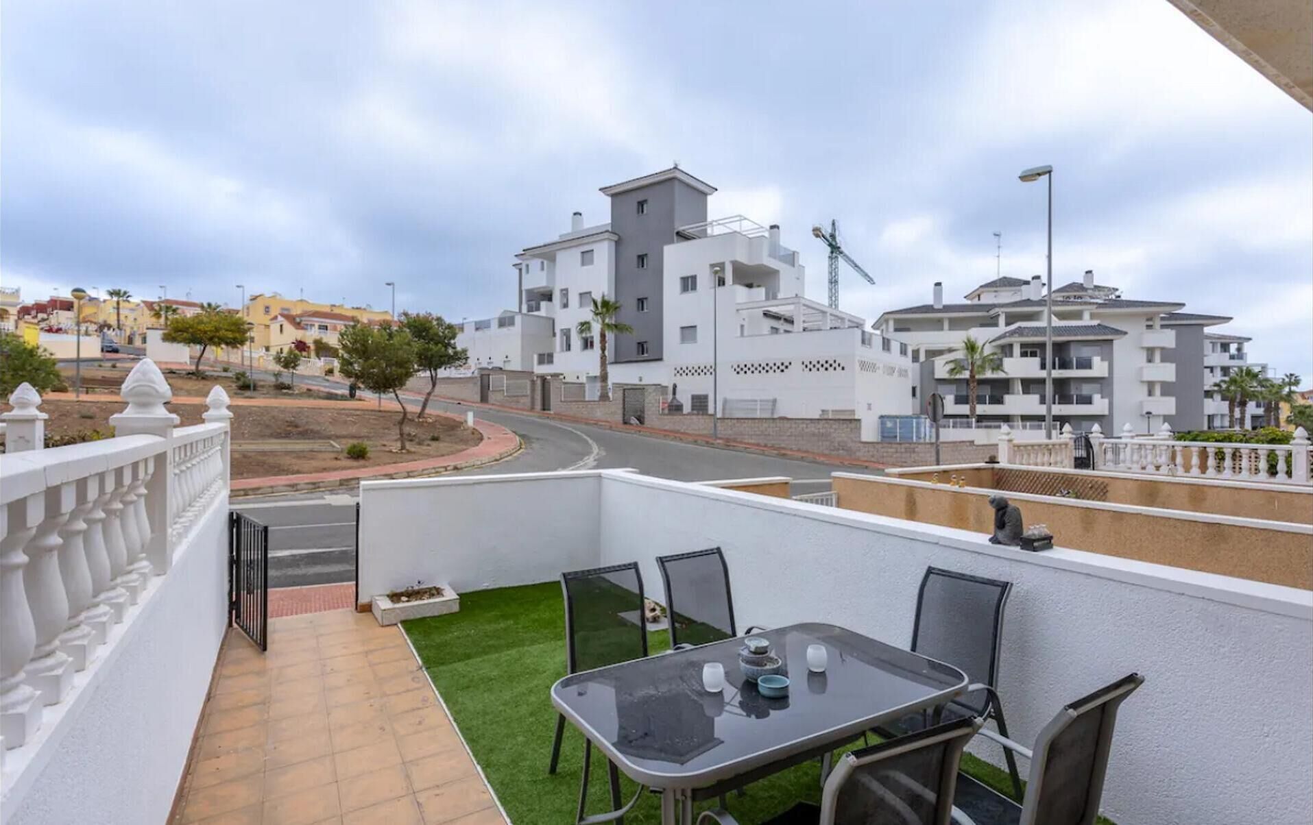 A balcony with a table and chairs, a view of a residential area with modern buildings, and a street in the background.