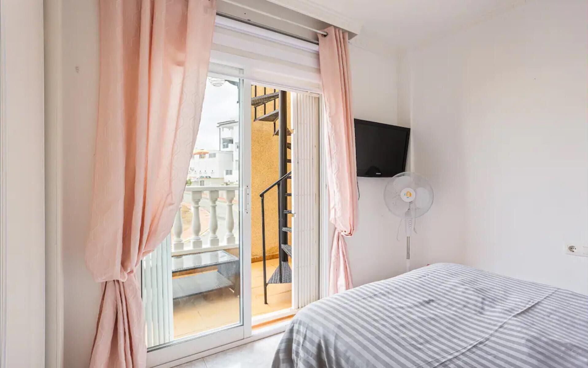A bedroom with a bed, a TV mounted on the wall, and a balcony with a railing.