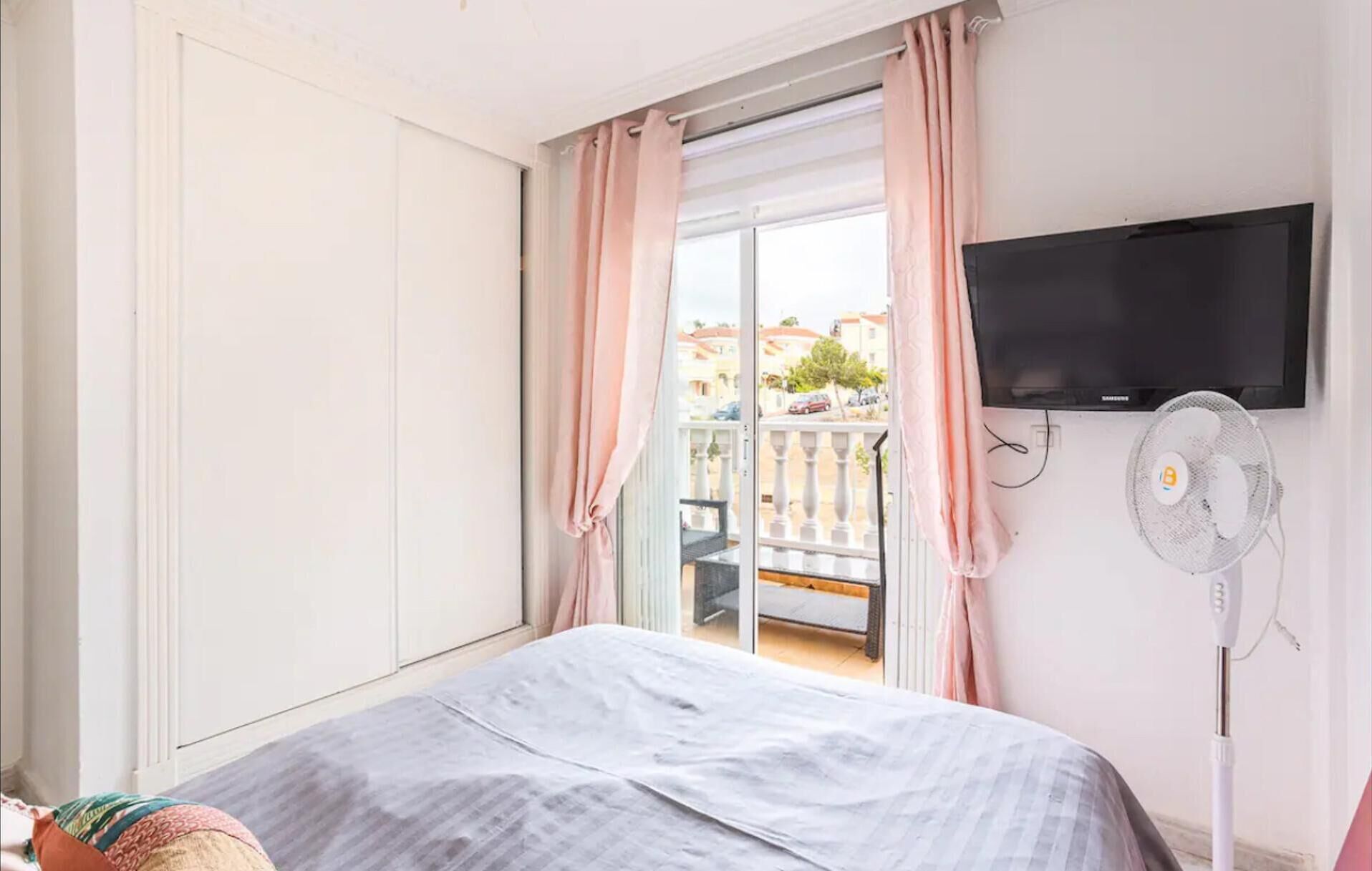 A bedroom with a bed, a TV, a fan, and a balcony with curtains.