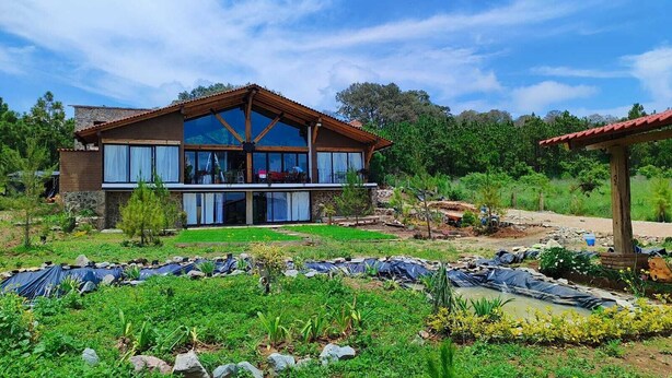 A two-story house with a large balcony, surrounded by a garden with various plants and a small pond. - Charming Chalet in Tapalpa, Mexico with Game Lounge & Pet-Friendly Amenities! (Tapalpa)