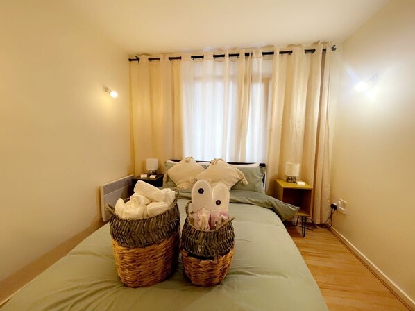 A hotel room with a bed, two baskets of towels, a bedside table with a lamp, and a window with curtains.