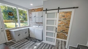 A kitchen with a brick wall, white cabinets, a sliding barn door, and a window with artwork. - IT’S FOOTBALL TIME AT THE HIDDEN TIGER! (Baton Rouge)