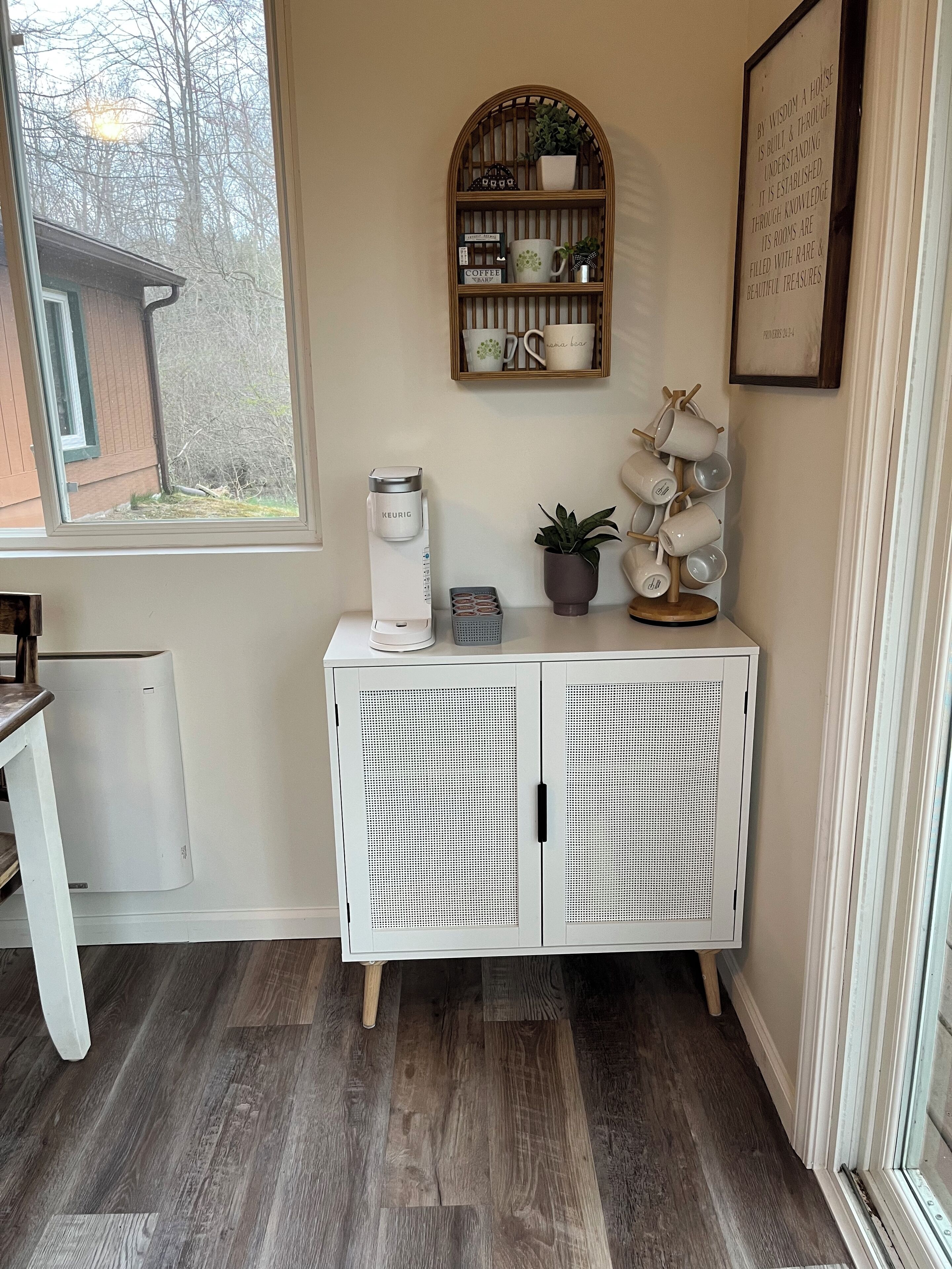 Eine Ecke eines Zimmers mit einem weißen Schrank, einer Keurig-Kaffeemaschine, einem kleinen Regal mit Tassen und einer Pflanze sowie einem Fenster mit Blick auf Bäume.