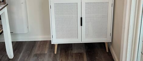 Eine Ecke eines Zimmers mit einem weißen Schrank, einer Keurig-Kaffeemaschine, einem kleinen Regal mit Tassen und einer Pflanze sowie einem Fenster mit Blick auf Bäume.