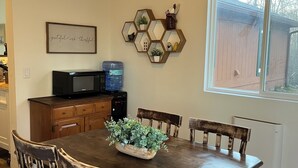 Zona pranzo con tavolo in legno, sedie, ventilatore a soffitto, forno a microonde, erogatore d'acqua e finestra con vista su una recinzione in legno.