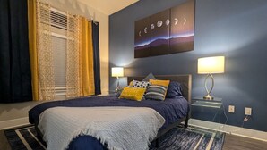 Ein Schlafzimmer mit einem Bett, zwei Nachttischen mit Lampen, einem Wandbild und einem Fenster mit Vorhängen.