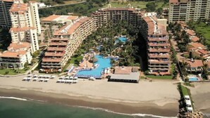 An aerial view of a resort with a large circular pool surrounded by multiple-story buildings, a beach area, and a rocky outcrop. - Regal 3bd PH with private pool in Beach Resort Spa (Marina Vallarta, Puerto Vallarta)