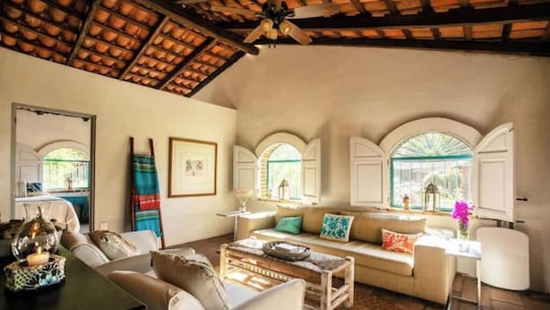 A cozy living room with a sofa, coffee table, and a ceiling fan. There are arched windows and a mirror on the wall.
